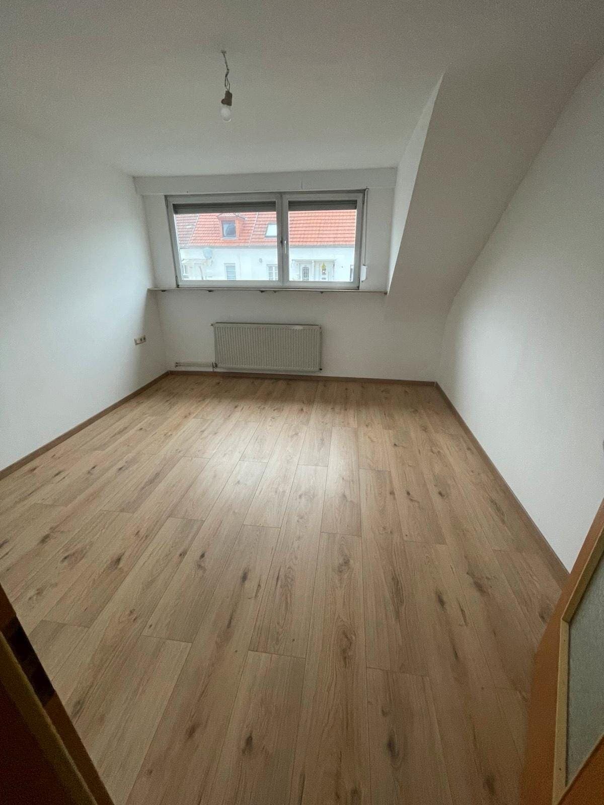 Fertiges Dachzimmer mit weißen Schrägen, neuem hellen Laminatboden und Heizkörper unter dem breiten Fenster