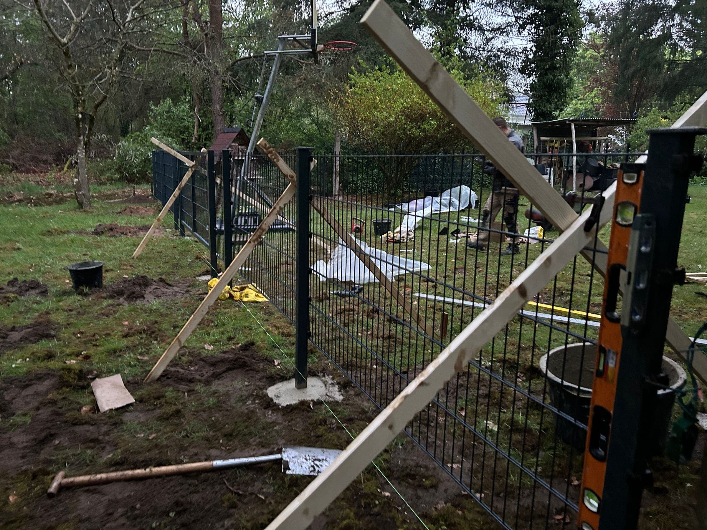 Montage eines anthrazitfarbenen Doppelstabmattenzauns: Pfosten mit Holzlatten abgestützt, ausgehobene Pfostenlöcher und eine Wasserwaage am Pfosten
