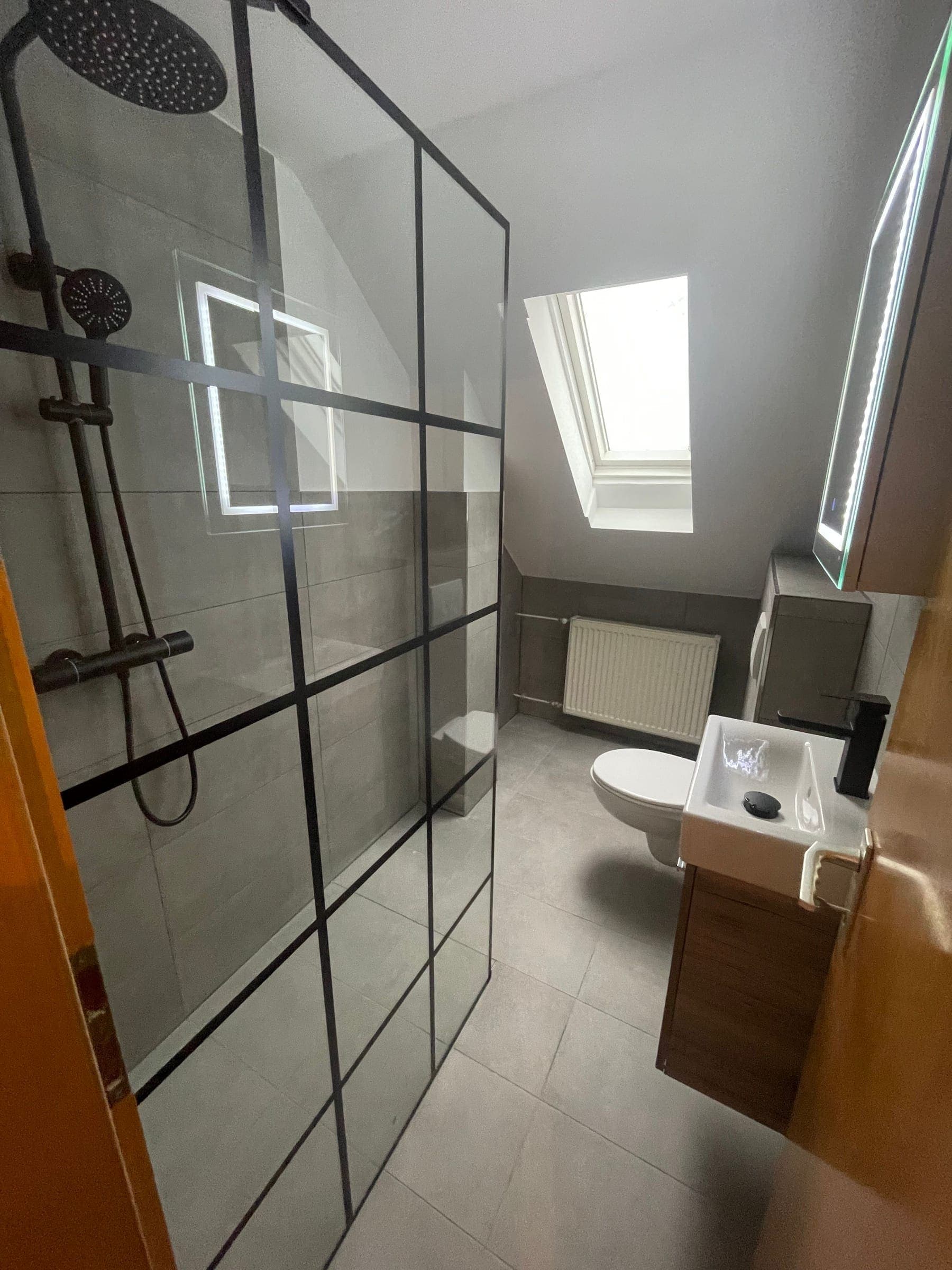 Fertiges Bad mit Walk-in-Dusche hinter schwarz gerasterter Glaswand, grauen Großformatfliesen und Waschtisch aus dunklem Holz