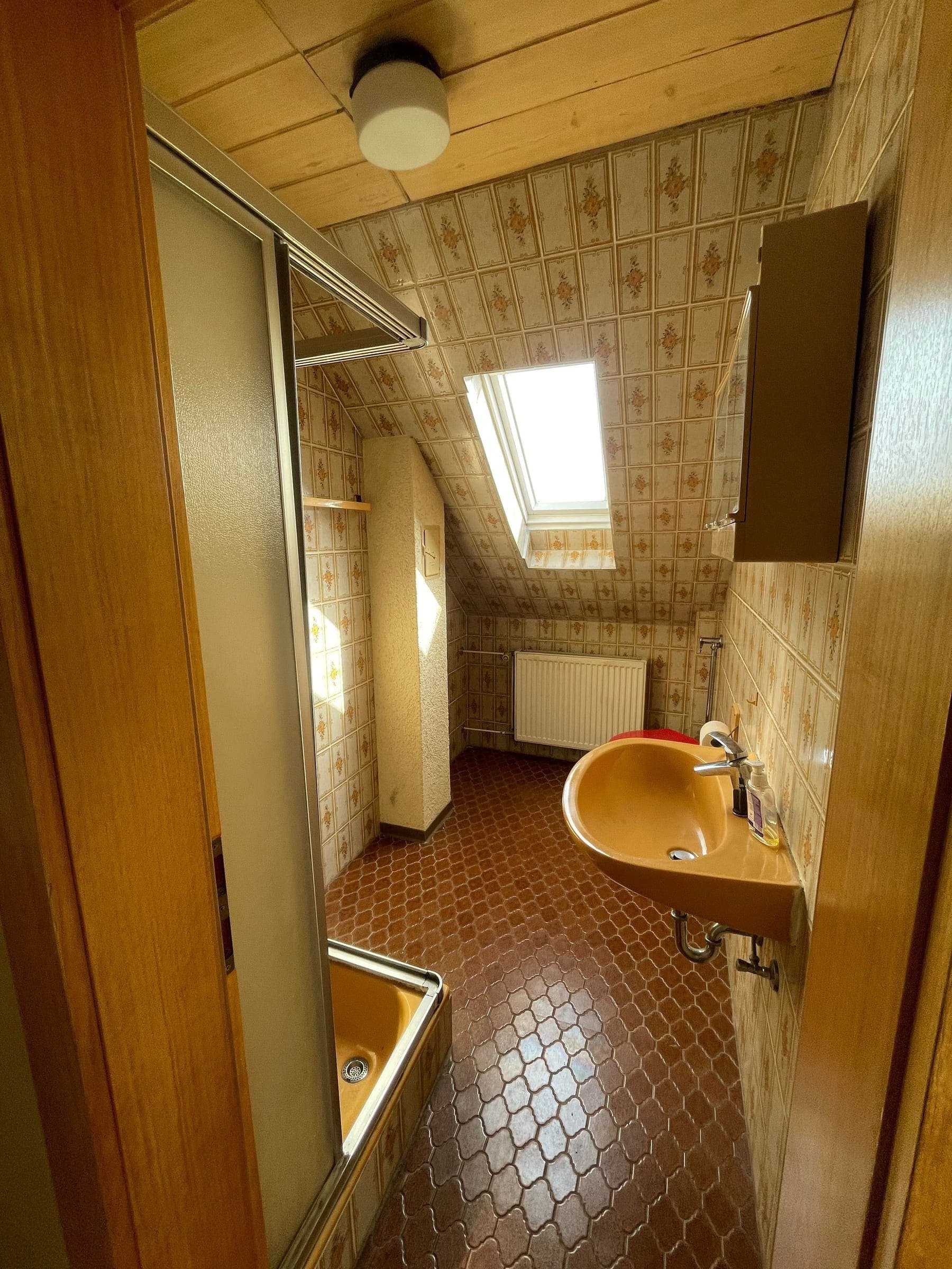 Dachbad aus den 1970er-Jahren mit gemusterten Wandfliesen, orangefarbenem Eckwaschbecken, orangefarbener Wanne hinter Schiebetür und Holzdecke